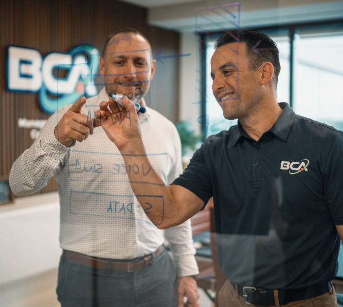 Two BCA team members brainstorming on a glass wall, smiling as they draw diagrams and notes in a modern office space.