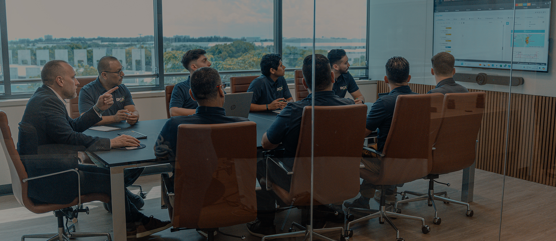 BCA IT team gathered in a modern glass-walled conference room, attentively viewing a presentation on a large screen. The team is seated around a table with laptops open, engaged in discussion, with a view of the cityscape through large windows in the background.