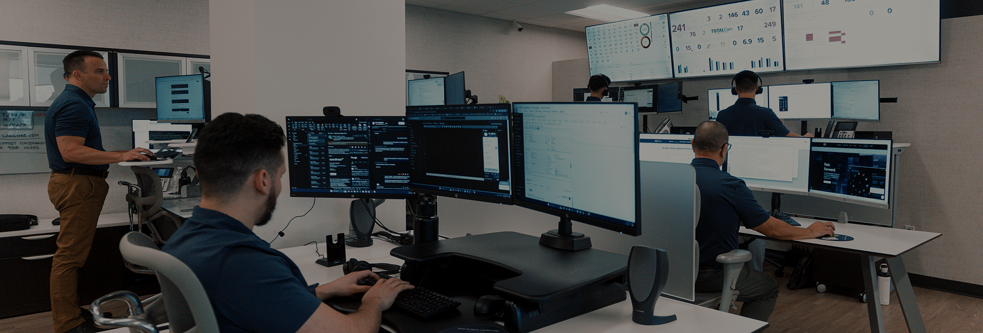 BCA IT team actively working in a modern operations center equipped with multiple monitors displaying dashboards, emails, and support systems. Employees are seated and standing at workstations, monitoring IT infrastructure and responding to issues in real-time. Large wall-mounted screens display key performance metrics and system health indicators.