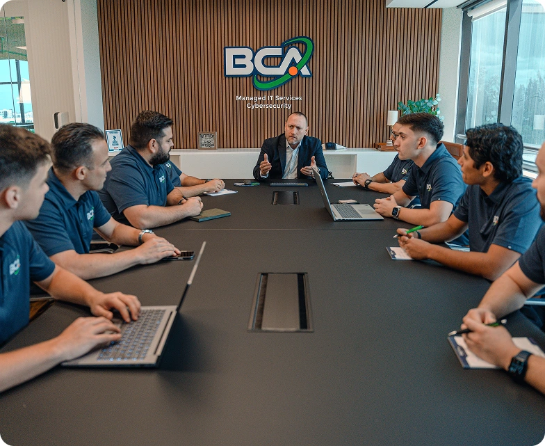 BCA team gathered around a conference table during a leadership meeting, with the company logo and “Managed IT Services Cybersecurity” displayed on the wall.