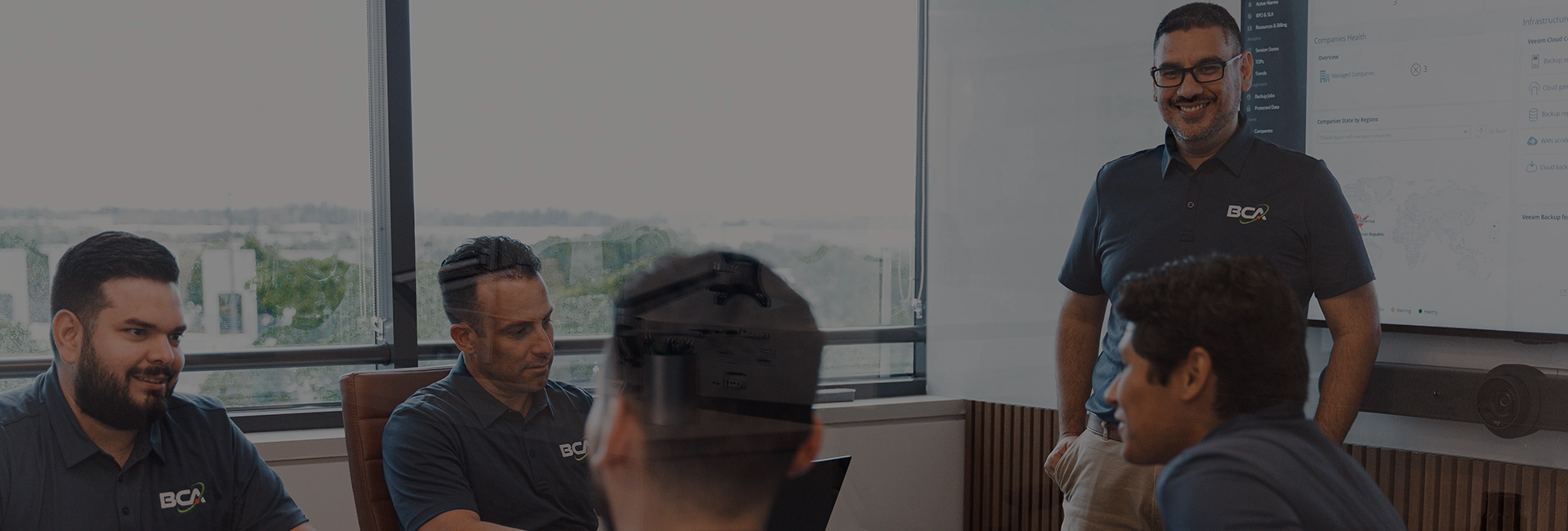 BCA IT team members in a meeting room, collaborating around a conference table. One team member stands in front of a screen displaying IT management dashboards, while others are seated, engaged in discussion. The room has large windows with natural light and a professional, modern atmosphere.