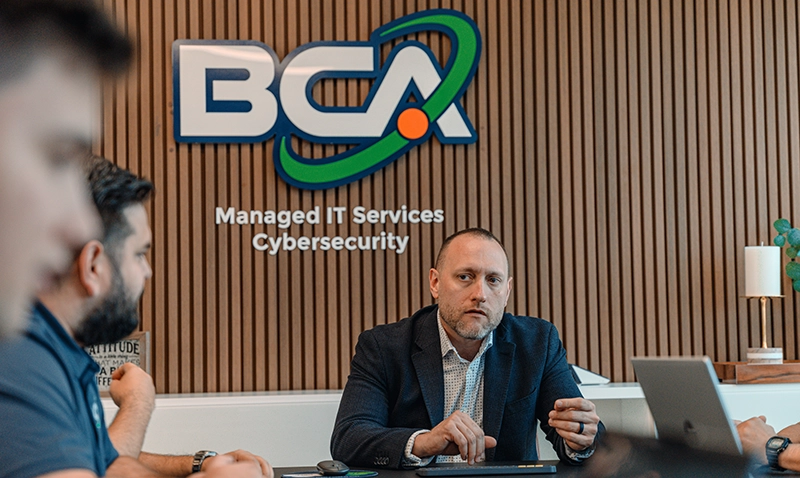 BCA executive leading a meeting with team members in a modern conference room, with a logo reading "Managed IT Services Cybersecurity" in the background.