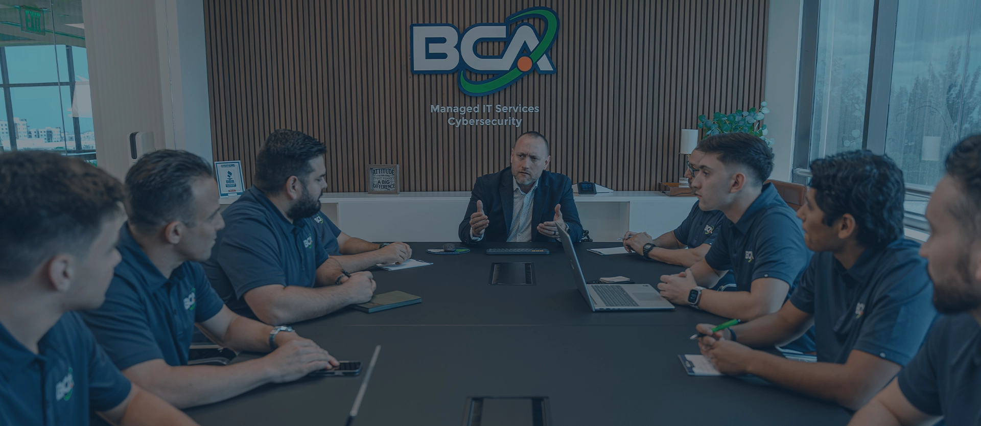 A manager sits at the head of a conference table discussing with IT staff, under a ‘BCA Managed IT Services Cybersecurity’ sign.