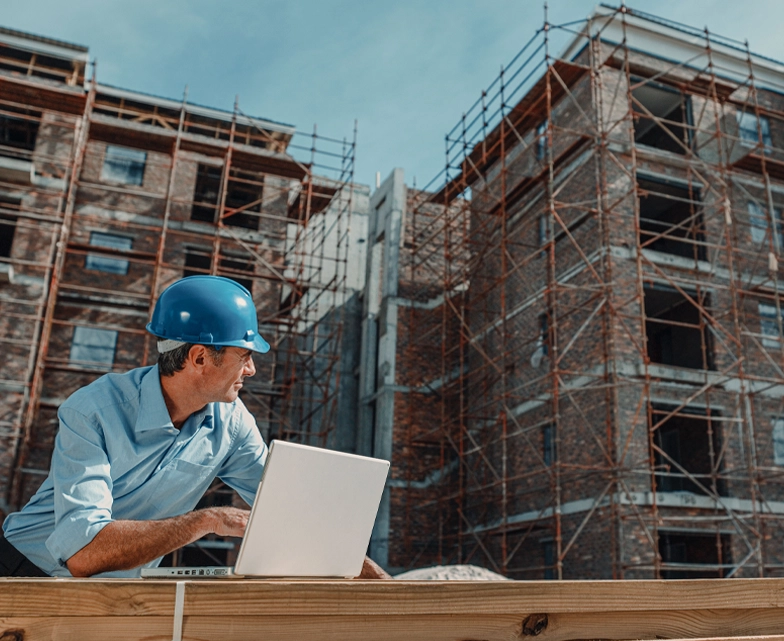 Construction professional using a laptop on-site with scaffolding and a multi-story building under construction in the background.