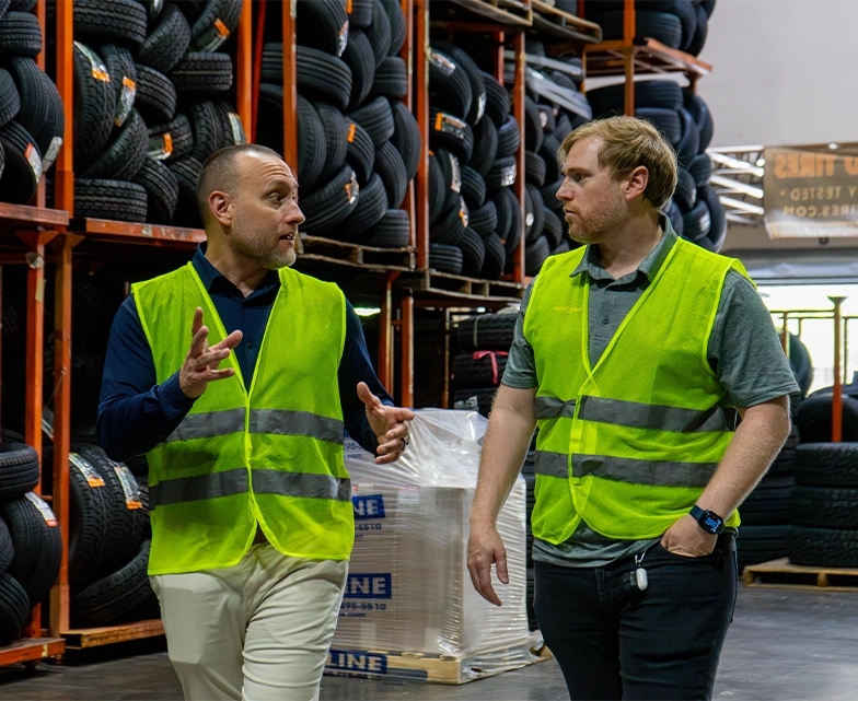 Two men in safety vests have a discussion while walking through a tire warehouse, highlighting an on-site client visit in an industrial setting.