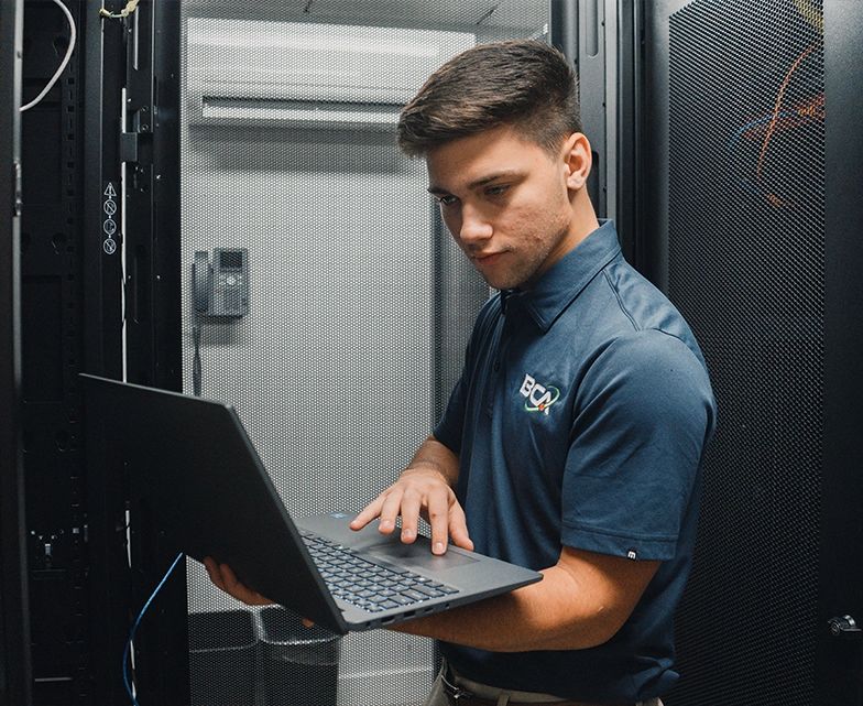 BCA IT engineer checking systems on a laptop inside a server rack room.