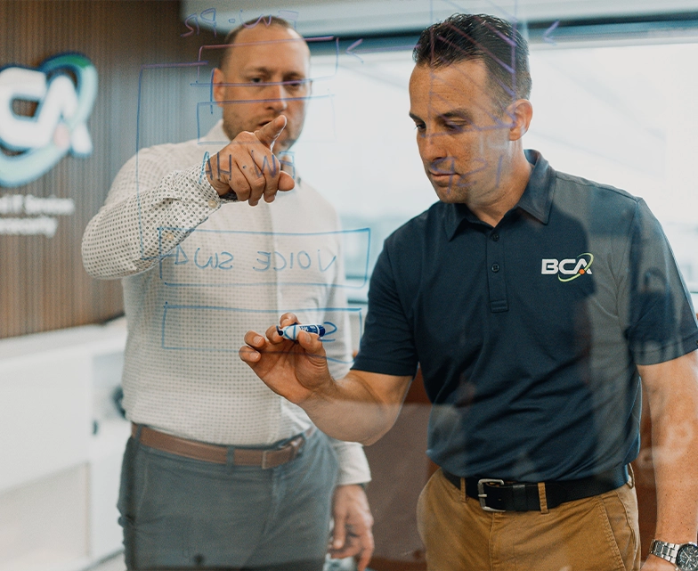 Two BCA team members collaborate on a technical diagram drawn on a glass board, mapping out IT infrastructure plans during a strategic planning session.