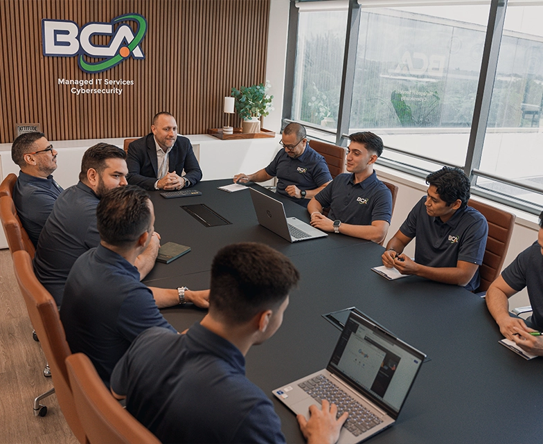 The BCA team gathers around a conference table for a collaborative meeting, with laptops and notebooks open as they discuss managed IT services and cybersecurity.
