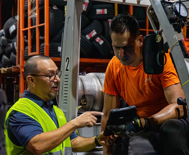 A BCA technician in a reflective vest points to a tablet while assisting a forklift operator inside a tire warehouse, representing IT support for manufacturing.