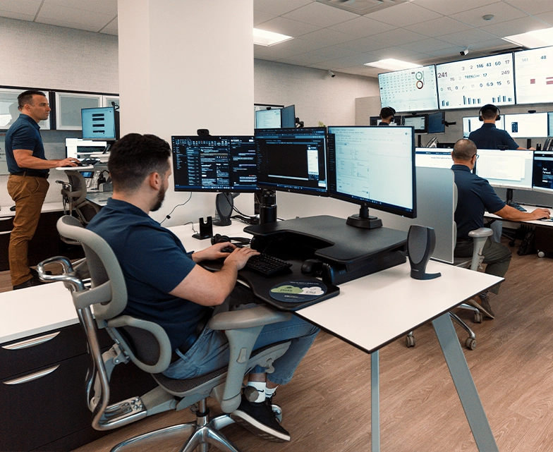 BCA technicians oversee real‑time dashboards on several monitors in a modern network operations center, providing continuous IT and cybersecurity monitoring.