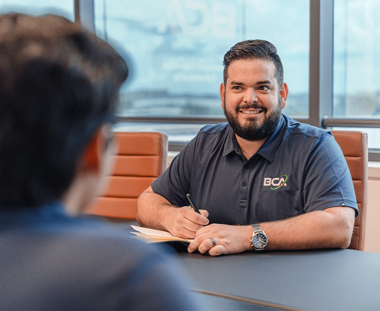 A smiling BCA team member takes notes during a meeting in a bright office, wearing a navy BCA-branded polo shirt.