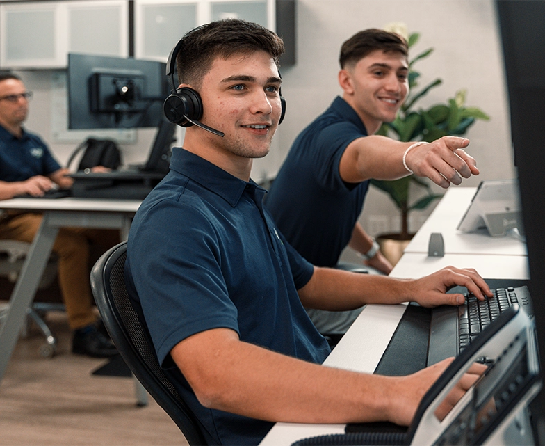 Two young BCA IT support specialists wearing headsets collaborate at a workstation, smiling as they assist clients in a modern office environment.