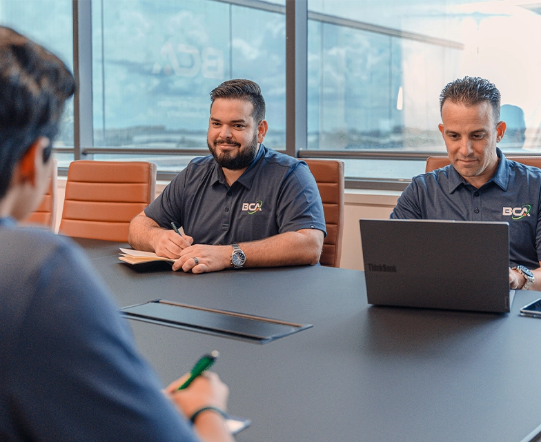 Two BCA team members participate in a meeting, one taking notes while the other works on a laptop, seated across from a colleague.