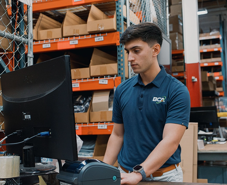 A BCA technician monitors operations on a desktop computer in a warehouse environment, surrounded by shelves stocked with cardboard boxes and IT equipment.