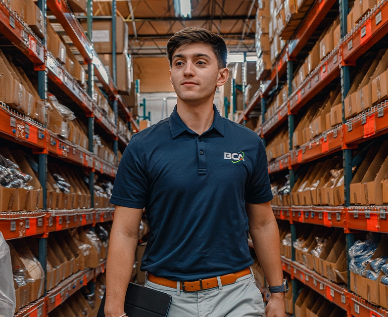 A BCA team member in a navy polo shirt walks confidently through a warehouse aisle lined with labeled inventory bins, carrying a tablet.
