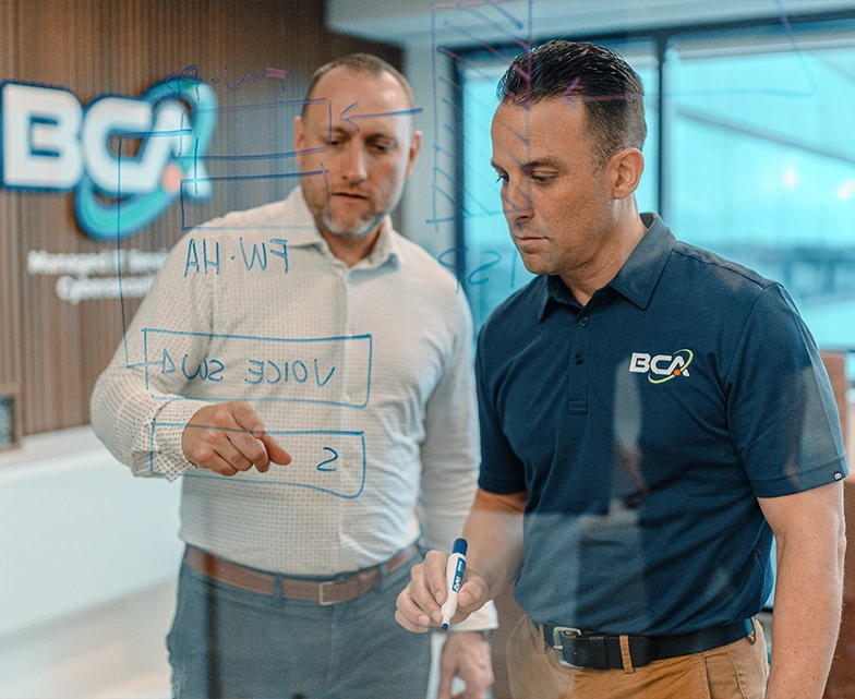Two BCA team members collaborate at a glass board, discussing and diagramming IT infrastructure plans during a strategy session.