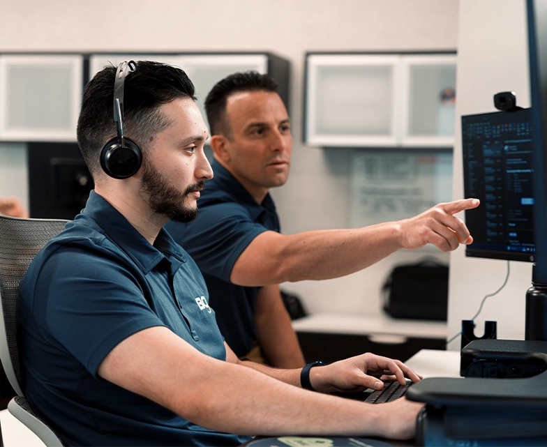 A BCA team member wearing a headset types on a keyboard as a colleague points to the screen, providing guidance during remote IT support.