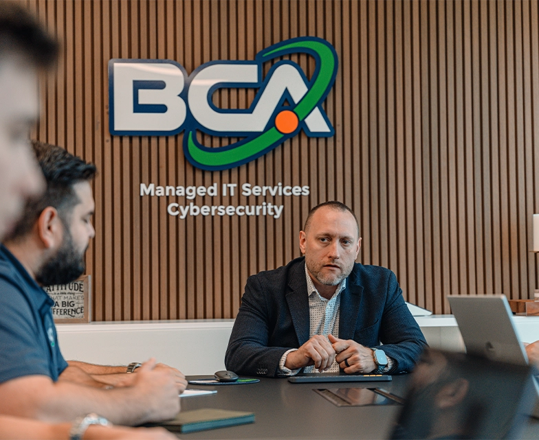 The BCA leadership team meets in the company conference room under a sign reading “Managed IT Services Cybersecurity,” discussing service improvements.