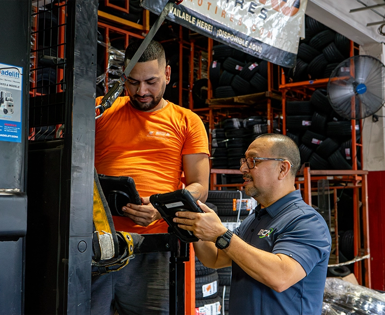 BCA technician providing on-site IT support to a warehouse worker using a tablet near a forklift, surrounded by stacked tires.