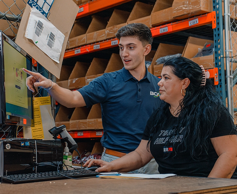 BCA technician assisting a warehouse employee with computer software at a workstation, with organized inventory shelves in the background.