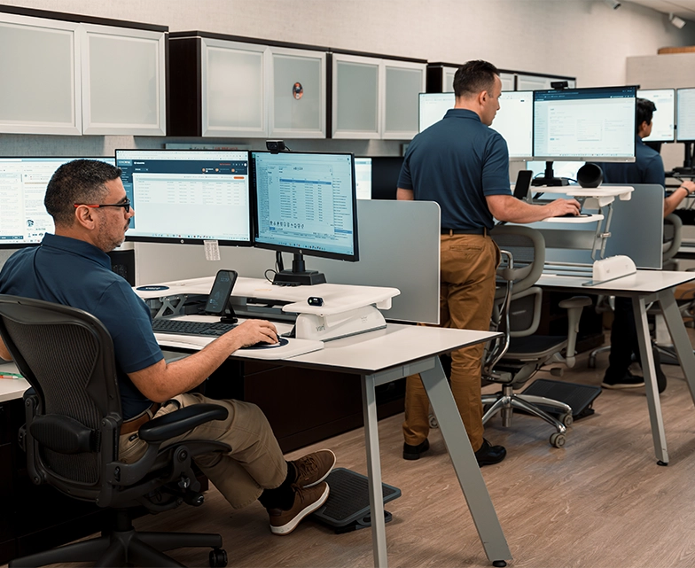 BCA team members work at sit-stand desks, monitoring client systems on multiple screens in a modern IT operations center.
