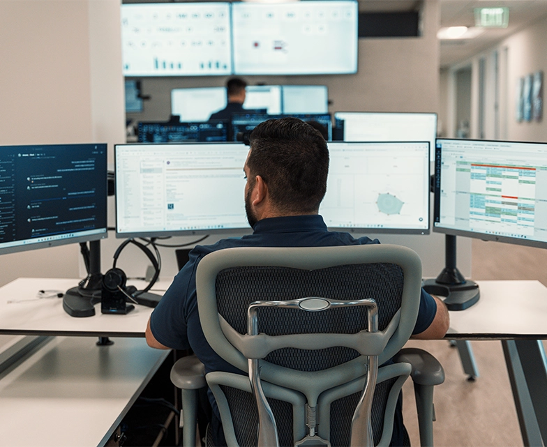 A BCA technician sits at a desk surrounded by five screens displaying dashboards, calendars, and alerts, overseeing IT systems and client communications.