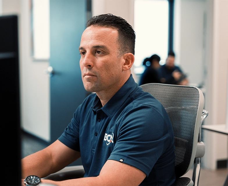 A focused BCA support technician in a navy polo works at his computer in a clean office space, handling remote IT help desk tasks.